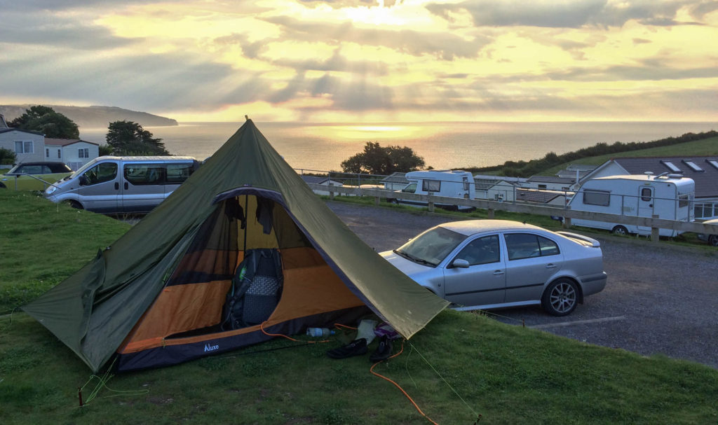 Ein Zelt steht auf einem Campingplatz. Im Hintergrund erkennt man den Sonnenuntergang über dem Meer.